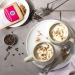 Two mugs of chai latte on white plate besides teaspoons, tea infuser, black peppercorns and sachet of drinking spice mix