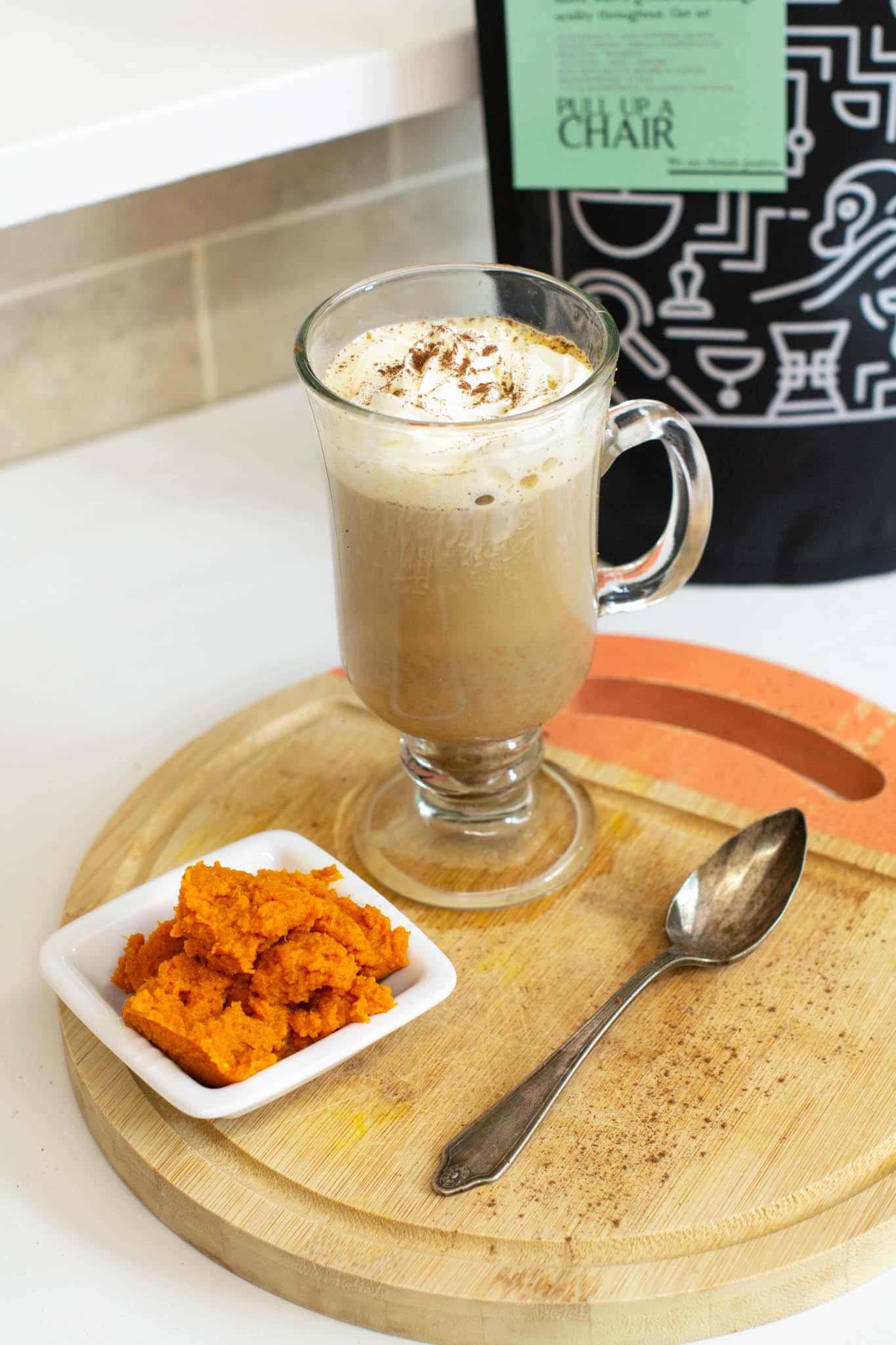 Pumpkin spice latte on circular chopping board with teaspoon, dish of pumpkin puree and Two Chimps Coffee bag
