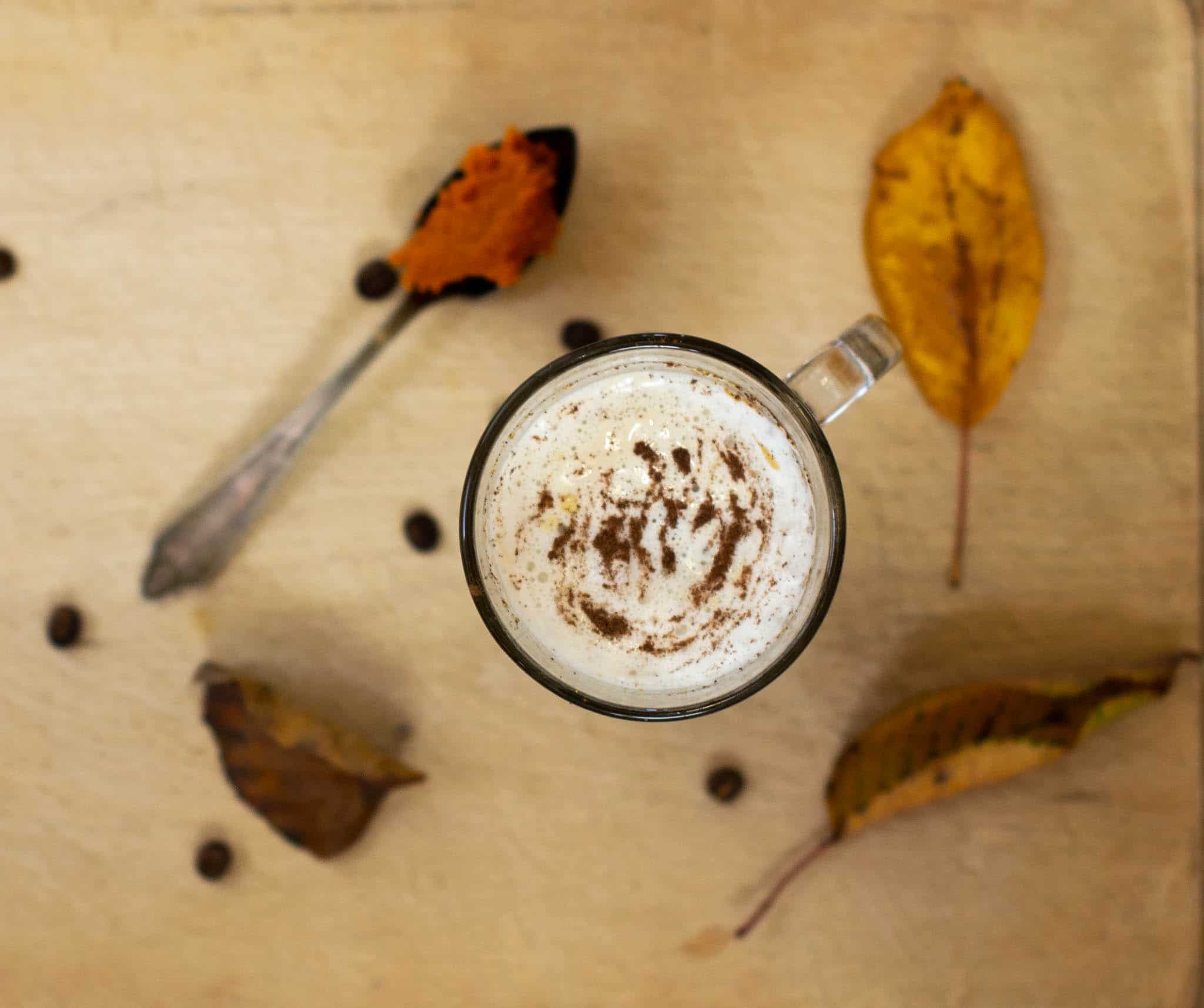 Latte glass from above surrounded by leaves, coffee beans and a teaspoon with pumpkin puree