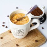 Coffee mug cake with teaspoon sitting on chopping board beside small teacup of coffee grounds