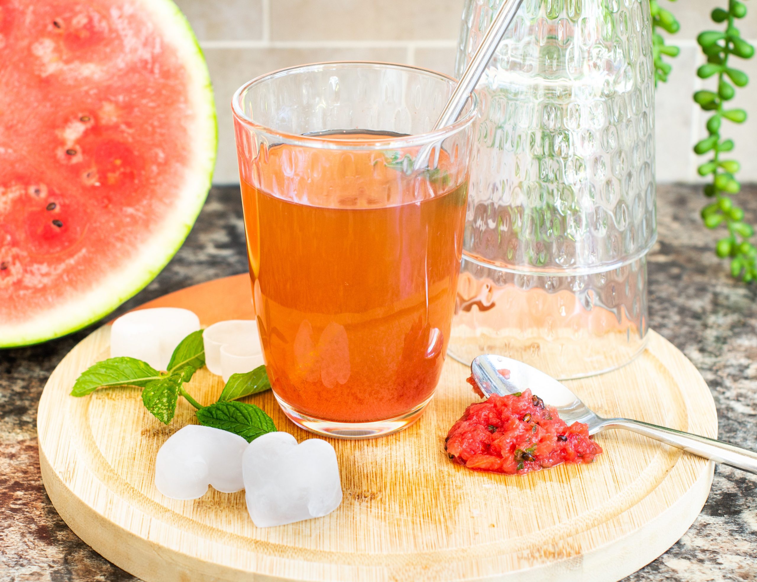 Glass of iced watermelon tea on a chopping board with ice, mint, empty glasses and watermelon flesh