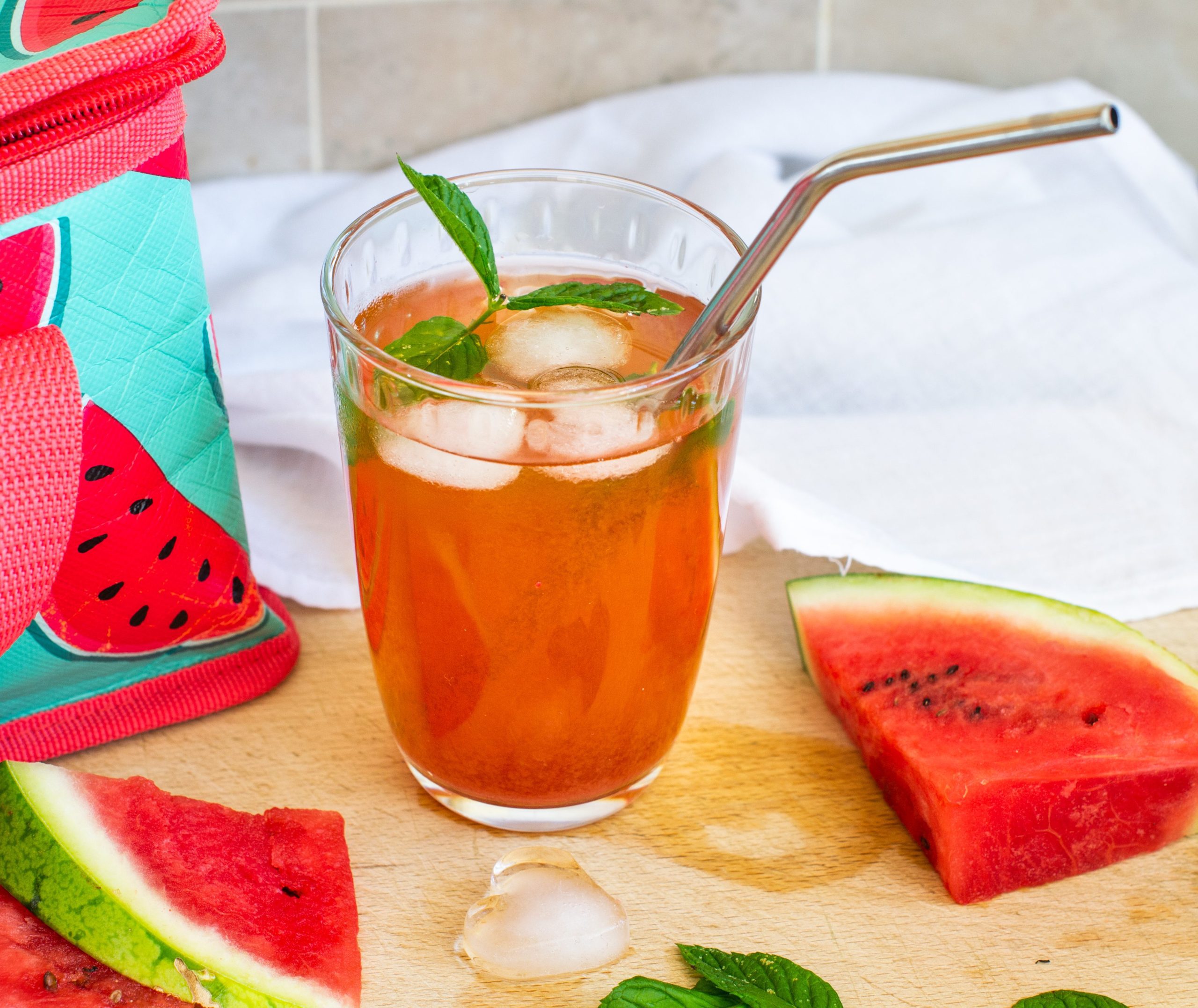 Glass of iced watermelon tea on a chopping board with ice, watermelon wedges and watermelon print lunchbag